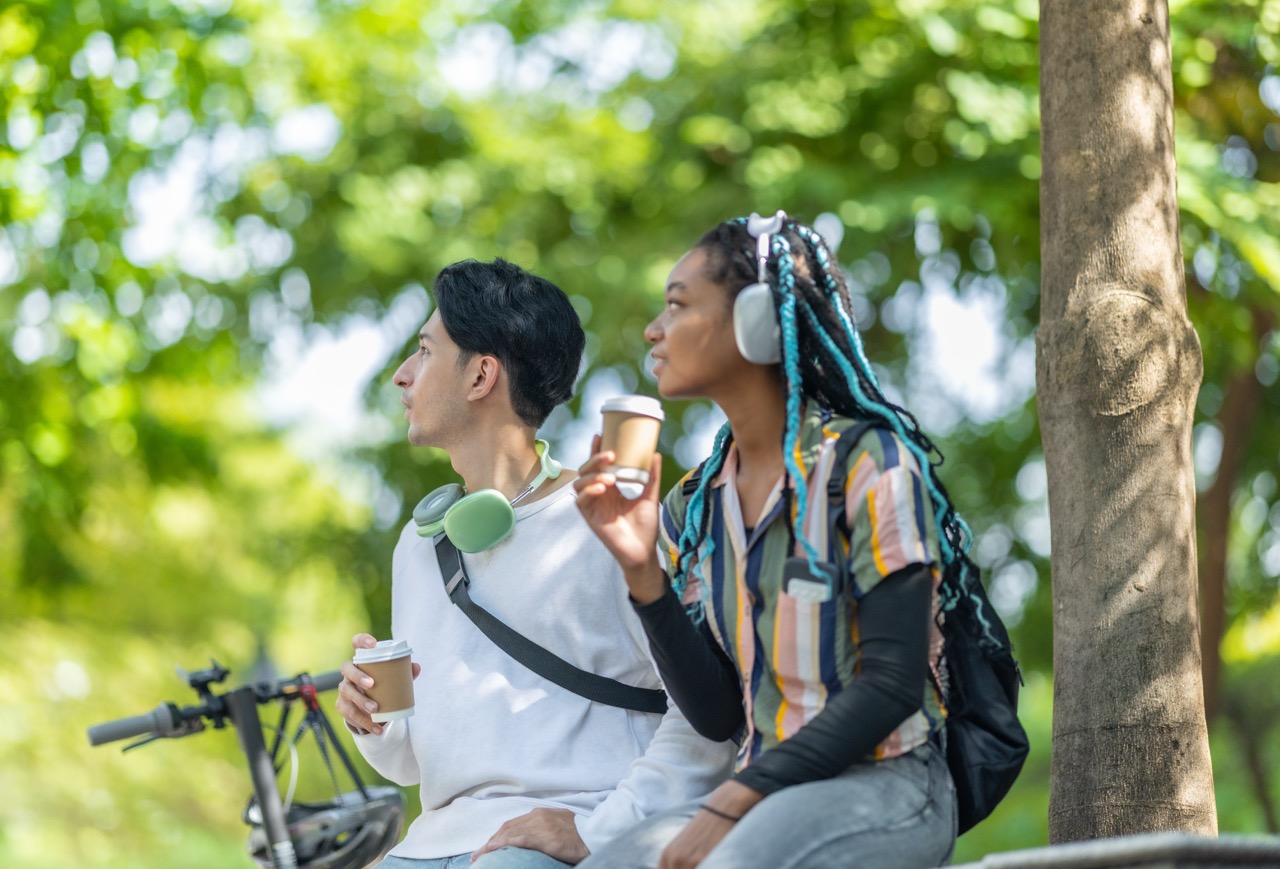 Friends enjoying coffee outdoors, wearing headphones, with a bicycle parked nearby and greenery in the background.